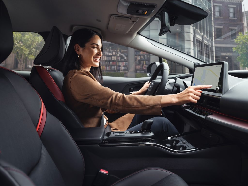 Woman sitting in the driver’s seat of a 2024 Toyota Prius Plug-in XSE, using the touchscreen navigation system inside a Black/Red SofTex interior.