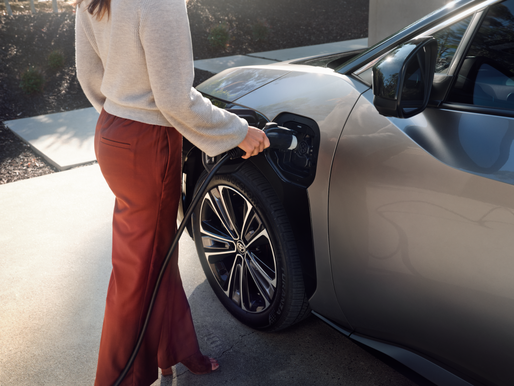 Woman charging a Toyota bZ4X electric vehicle in a residential driveway, close-up on the charging port and wheel.